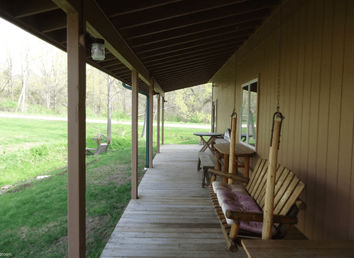A wooden porch with swings and a table, surrounded by greenery and a view of a road in the distance.