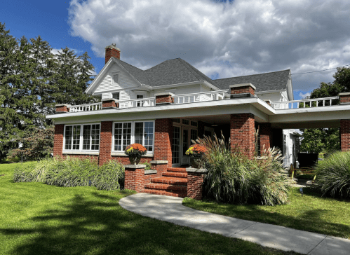 A large brick house with a porch, surrounded by greenery and under a partly cloudy sky.