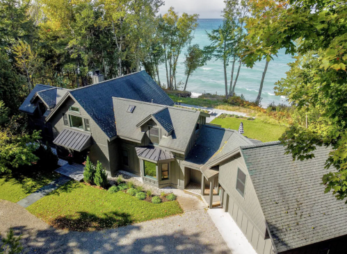 Aerial view of a modern house surrounded by trees, overlooking a blue lake and sandy shore.