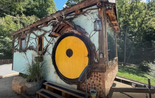 A whimsical tiny house with a large yellow circular door, surrounded by trees and natural elements.