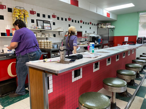 Two staff members work behind a diner counter with stools, preparing food in a retro-style kitchen.