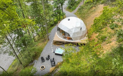 Aerial view of a geodesic dome cabin surrounded by trees, with a hot tub and fire pit nearby on a gravel path.