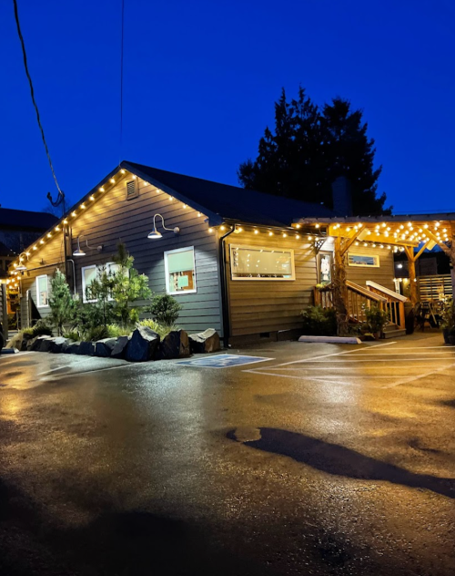 A cozy wooden building illuminated by string lights at dusk, with a wet pavement reflecting the lights.
