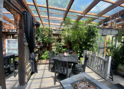 A cozy outdoor dining area with wooden tables, surrounded by greenery and a glass roof, creating a serene atmosphere.