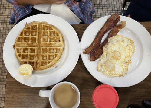 A plate with a waffle, two fried eggs, crispy bacon, a butter pat, and a cup of coffee on a wooden table.