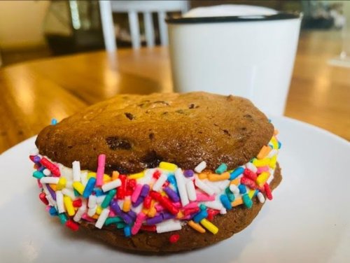 A chocolate chip cookie sandwich filled with colorful sprinkles, next to a white mug on a wooden table.