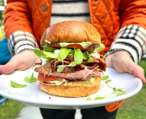 A person holds a large sandwich stacked with meats, tomatoes, and greens on a plate, wearing an orange quilted jacket.