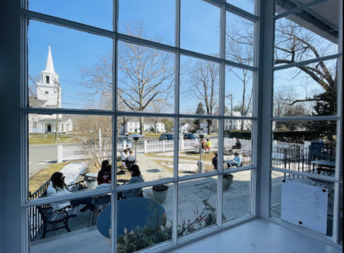 View through a window showing a sunny street scene with people sitting outside and a church steeple in the background.