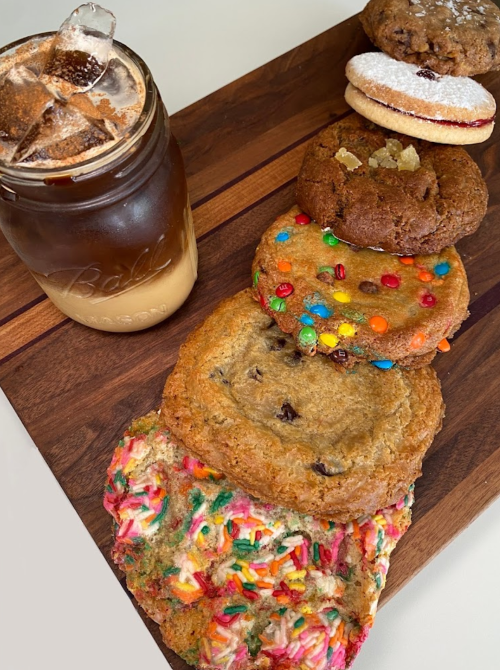 A wooden board displays a variety of colorful cookies alongside a jar of iced coffee with ice cubes.