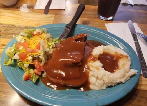 A blue plate with sliced meat in gravy, mashed potatoes, and a side salad with lettuce, tomatoes, and cheese.