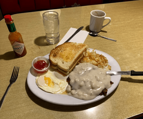 A plate with toast, hash browns, an egg, and chicken smothered in gravy, with a glass of water and hot sauce on the side.