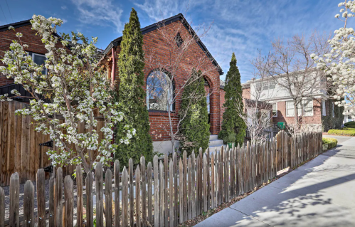 A charming brick house surrounded by trees and blooming flowers, with a wooden fence in front and a clear blue sky.