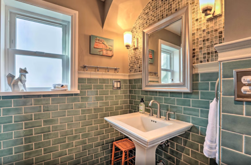 A stylish bathroom featuring a pedestal sink, green tiled walls, a mirror, and a small wooden stool.