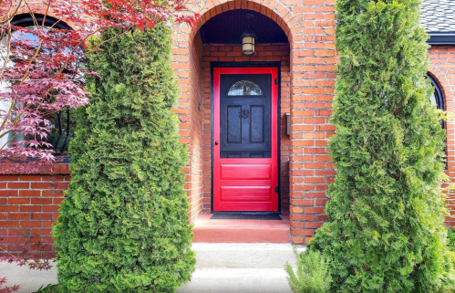 A red door framed by two tall green shrubs, set against a brick wall and archway.