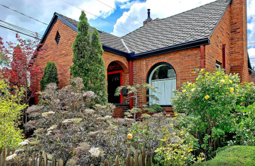 A charming brick house surrounded by lush greenery and colorful flowers under a partly cloudy sky.