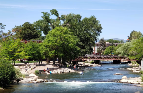A serene river scene with lush trees, a bridge, and people enjoying the outdoors on a sunny day.