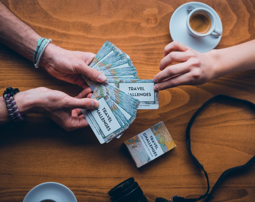 Two hands exchanging travel challenge cards over a wooden table, with a cup of coffee and a camera nearby.
