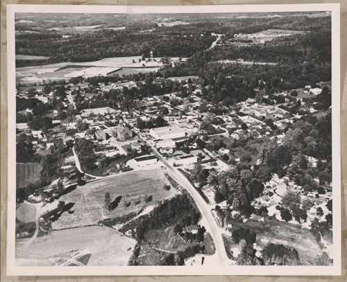 Aerial view of a small town surrounded by fields and forests, featuring roads and buildings in a rural landscape.