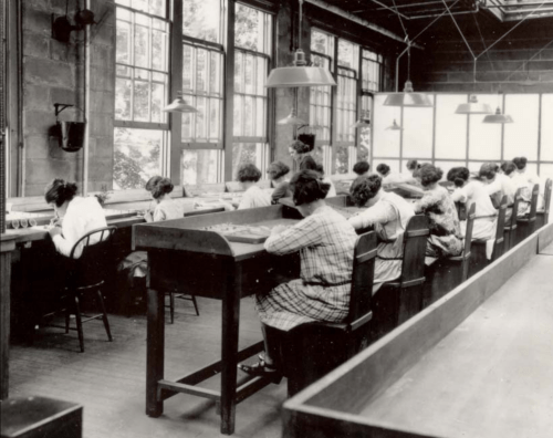 Black and white photo of women working at long tables in a well-lit room with large windows.