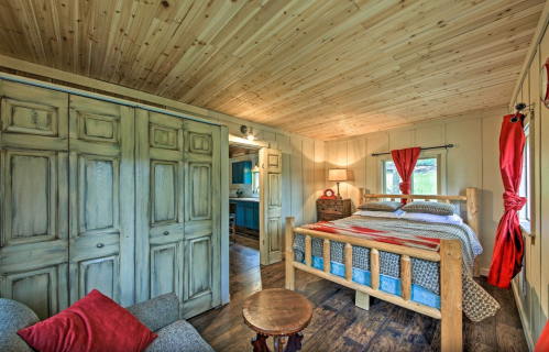 Cozy bedroom with wooden walls, a log bed, red curtains, and a small table, featuring a rustic, inviting atmosphere.