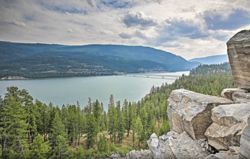 Scenic view of a river surrounded by mountains and forests under a cloudy sky. Large rocks in the foreground.
