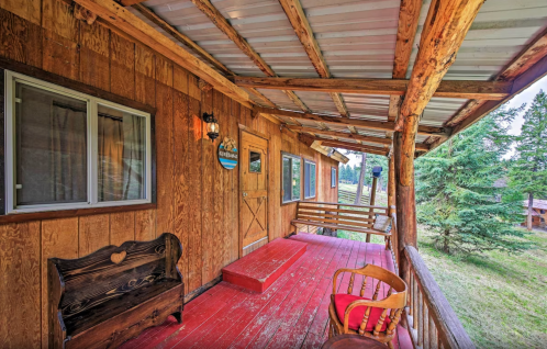 A rustic wooden porch with a red floor, featuring a bench and a chair, surrounded by greenery and trees.