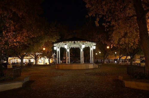 A lit gazebo in a park at night, surrounded by trees with autumn leaves and soft street lighting.