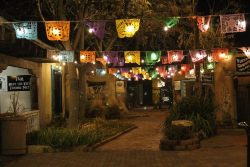 Colorful papel picado banners hang above a dimly lit pathway in a festive outdoor setting.