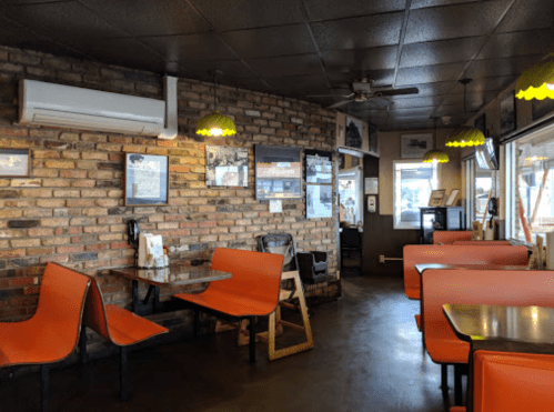 Interior of a cozy restaurant featuring brick walls, orange chairs, and warm lighting. Tables are set for dining.