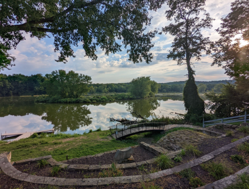 A serene river scene with trees, a small bridge, and reflections on the water under a partly cloudy sky.