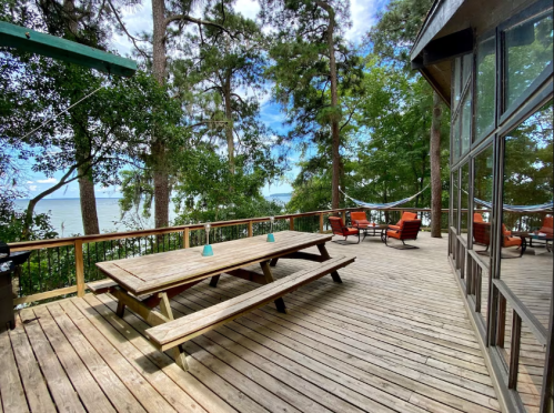 A wooden deck with picnic tables and chairs, surrounded by trees and overlooking a lake under a blue sky.