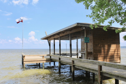 A wooden dock with a small shelter extends over calm water, featuring a flag flying in the breeze under a blue sky.