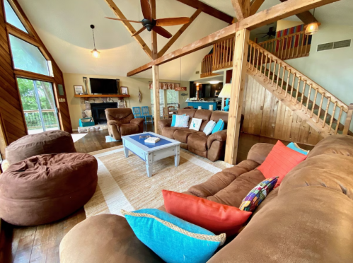 Cozy living room with brown sofas, colorful cushions, wooden beams, and a staircase, featuring a large window and a fireplace.