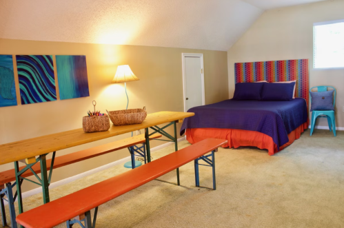 Cozy room with a bed, colorful artwork, a wooden table, and benches, featuring warm lighting and a bright atmosphere.