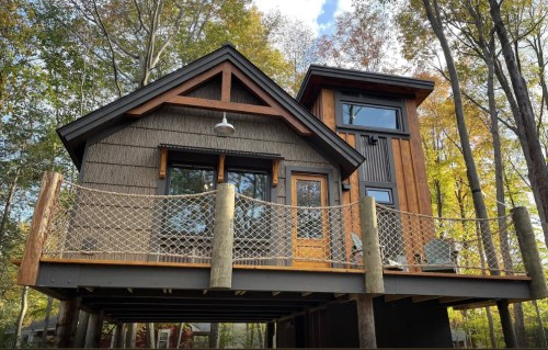 A modern cabin with a wooden deck, surrounded by trees, featuring large windows and a unique architectural design.