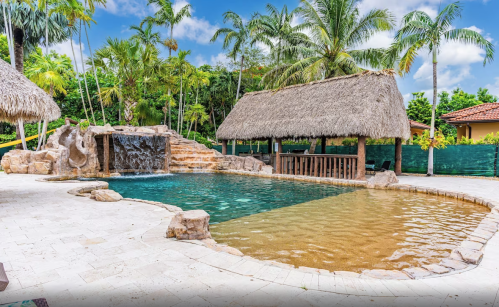 Tropical pool area with a waterfall, palm trees, and thatched-roof cabana under a blue sky.