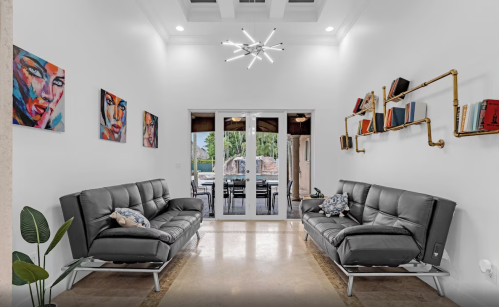Modern living room with two black sofas, colorful artwork, and a stylish light fixture, leading to an outdoor view.