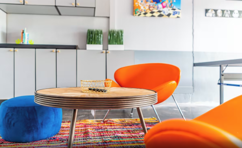 A modern living space featuring a round wooden table, orange chairs, a blue pouf, and colorful rugs.