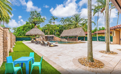 Tropical pool area with palm trees, thatched huts, lounge chairs, and colorful outdoor furniture on a sunny day.