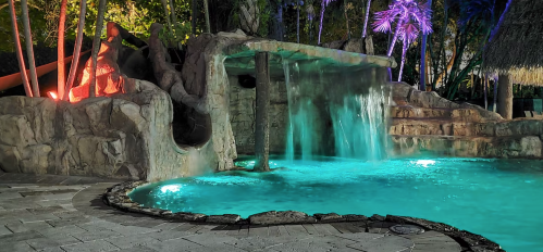 A tropical pool area featuring a waterfall, stone slide, and vibrant lighting surrounded by lush greenery.