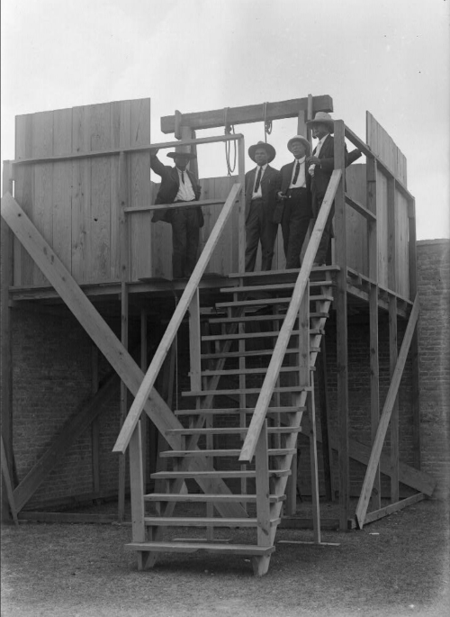 Three men in suits stand on a wooden platform with a gallows, featuring a staircase leading up to it.