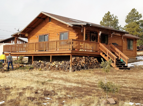 A wooden cabin with a large deck, surrounded by grass and stacked firewood, set against a cloudy sky.