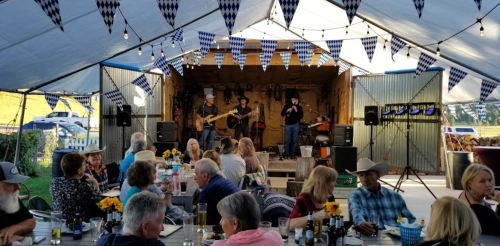 A lively outdoor event with a band performing on stage, surrounded by guests enjoying food and drinks under festive decorations.