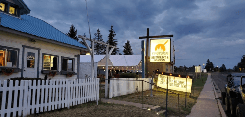 A restaurant and saloon with a sign, surrounded by trees and a white picket fence, under a cloudy sky.