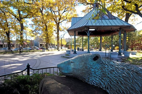 A bronze bear sculpture in a park with a gazebo and autumn trees in the background.