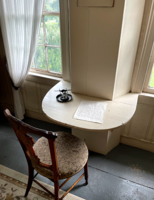 A small, curved wooden table by a window with a chair and a teapot on it, surrounded by light curtains.