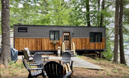 A modern tiny house with a wooden exterior, surrounded by trees and a gravel path, featuring outdoor seating.