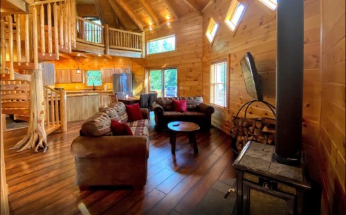 Cozy wooden cabin interior with a living area, fireplace, and open kitchen, featuring large windows and rustic decor.