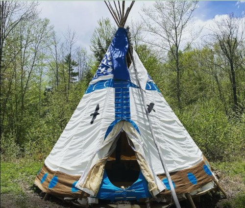 A colorful teepee with blue and white patterns, set in a lush green forest under a partly cloudy sky.