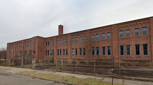 A large, abandoned brick building with multiple windows, surrounded by a chain-link fence and overgrown grass.